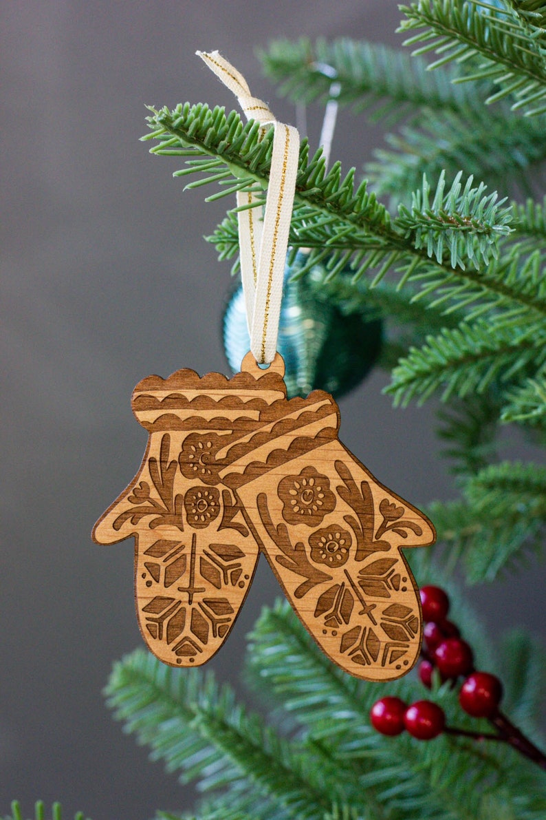 May include: A wooden Christmas ornament shaped like a pair of mittens with a floral design. The ornament is hanging on a Christmas tree with red berries.
