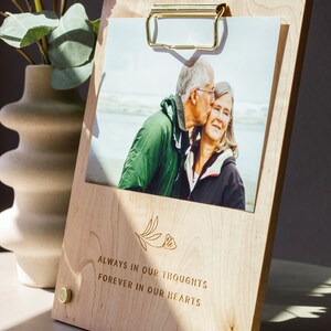 May include: A wooden photo frame with a gold clip and brass accents. The frame holds a photo of an older couple. The frame is engraved with the text "Always in our thoughts, forever in our hearts".