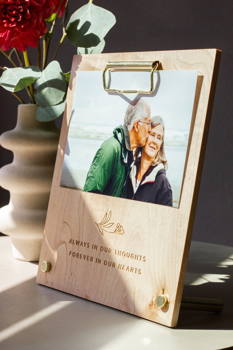 May include: A wooden photo frame with a gold clip and brass accents. The frame holds a photo of an older couple. The frame is engraved with the text "Always in our thoughts, forever in our hearts".