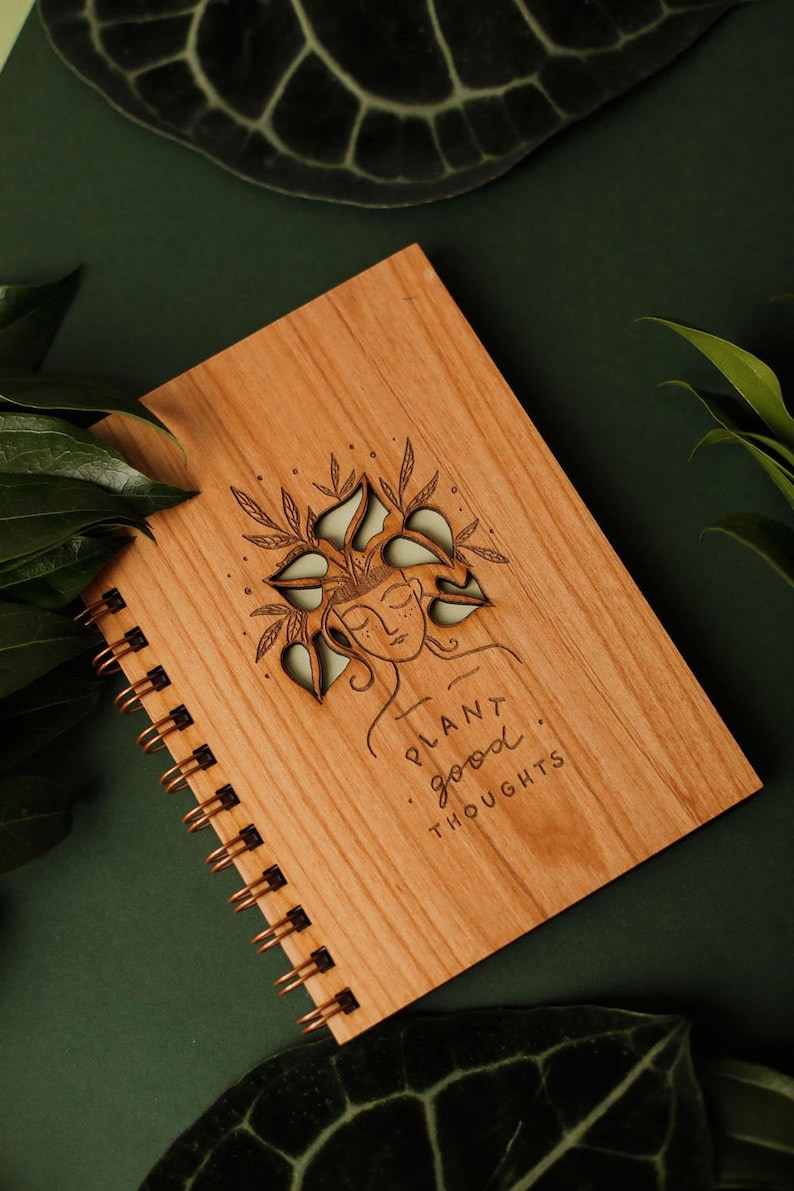 May include: Wooden spiral-bound notebook with a laser-etched design. The cover features a woman's face with leaves in her hair and the words "Plant good thoughts." The notebook has a copper-colored spiral binding.