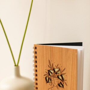 May include: A wooden spiral-bound notebook with a laser-cut design of a person with plants growing from their head. The text "PLANT good THOUGHTS" is below the illustration. The notebook is open, revealing white pages.