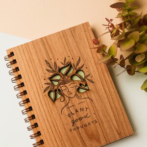 May include: Wooden spiral-bound notebook with a laser-cut design of a woman with plants in her hair. The notebook cover is engraved with the words "PLANT good THOUGHTS". The notebook is brown with a copper-colored spiral binding.