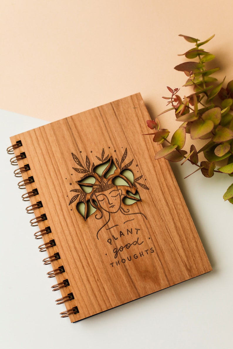 May include: Wooden spiral-bound notebook with a laser-cut design of a woman with plants in her hair. The notebook cover is engraved with the words "PLANT good THOUGHTS". The notebook is brown with a copper-colored spiral binding.