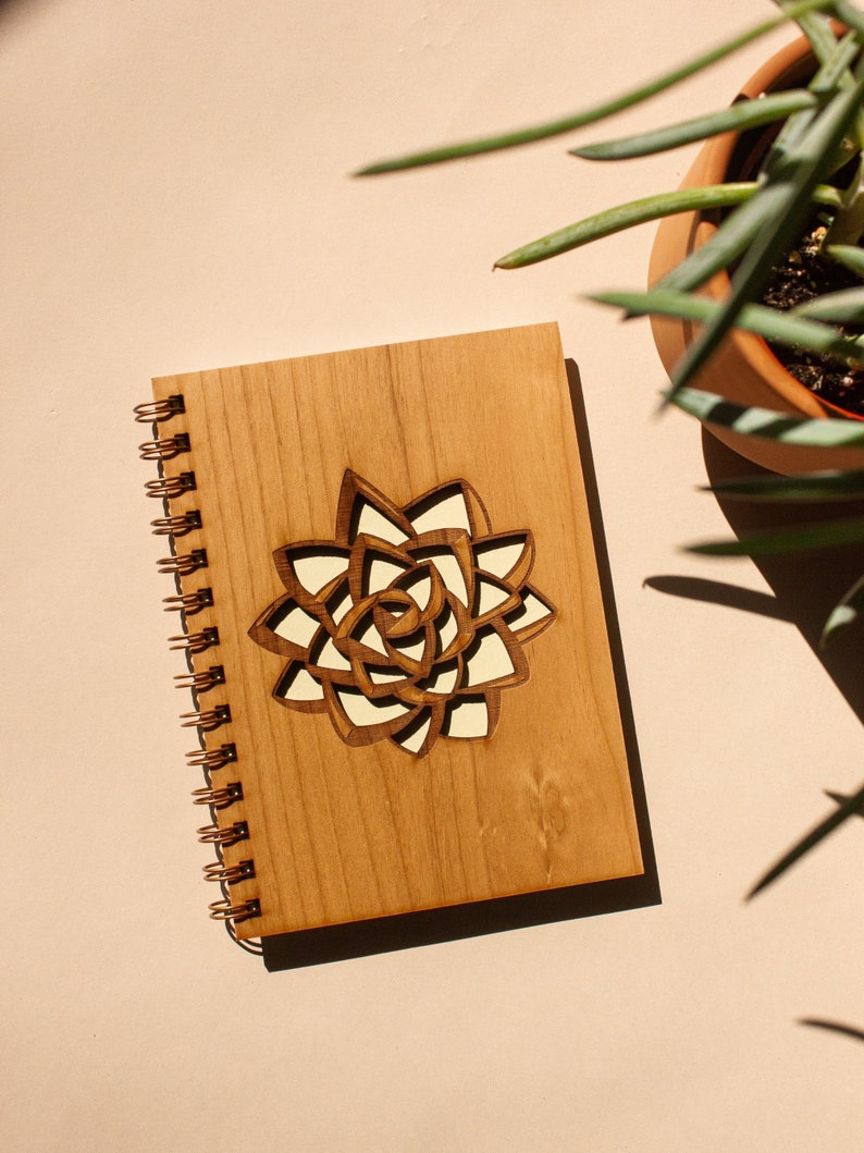Puede incluir: Un cuaderno de madera con un dise&ntilde;o de suculenta cortado con l&aacute;ser en la portada. El cuaderno tiene una encuadernaci&oacute;n en espiral y un acabado de madera marr&oacute;n claro. El dise&ntilde;o de la suculenta tiene un fondo crema.