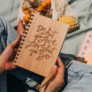 May include: A wooden journal with the words "Do what you love, love what you do" engraved on the cover. The journal is open and a person is holding a gold pen.