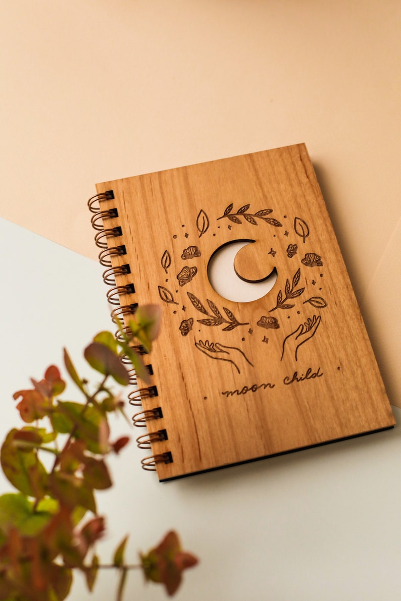 May include: A wooden journal with a spiral binding. The cover features a crescent moon cutout surrounded by leaves, stars, and hands reaching up. The text "moon child" is engraved below the design.