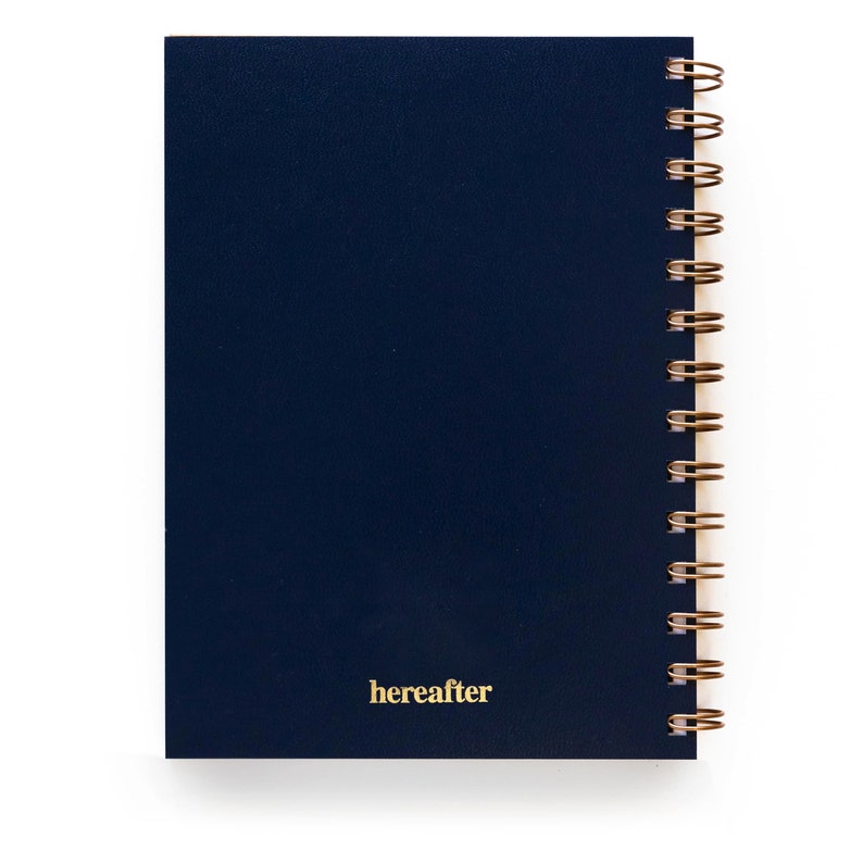 Puede incluir: Un cuaderno azul marino con encuadernaci&oacute;n en espiral de alambre dorado. La palabra "hereafter" est&aacute; impresa en dorado en la esquina inferior derecha de la portada. El cuaderno est&aacute; sobre un fondo blanco.