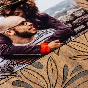 May include: A close-up of a photograph showing a couple embracing. The woman is wearing a black leather jacket and the man is wearing a grey and white sweater. The photograph is on a wooden surface with a carved leaf pattern.