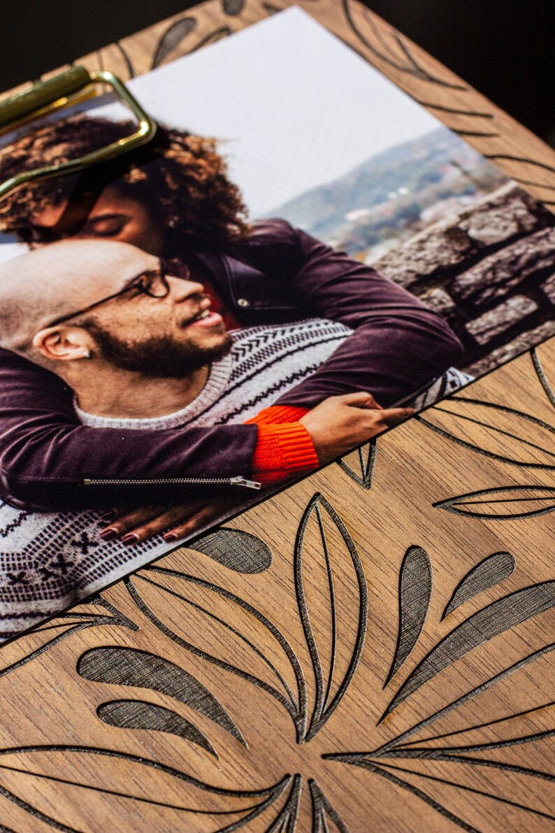 May include: A close-up of a photograph showing a couple embracing. The woman is wearing a black leather jacket and the man is wearing a grey and white sweater. The photograph is on a wooden surface with a carved leaf pattern.