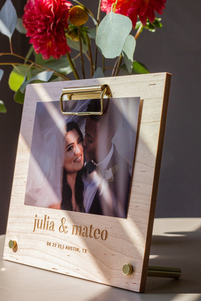 May include: A wooden photo frame with a gold clip, holding a wedding photo of a couple. The frame has the text "julia & mateo" and "06 22 21 | AUSTIN, TX" engraved on it.