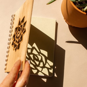 Puede incluir: Un cuaderno de madera con espiral y un dise&ntilde;o floral cortado con l&aacute;ser en la portada. El cuaderno est&aacute; abierto, revelando p&aacute;ginas en blanco. La sombra del dise&ntilde;o floral se proyecta sobre las p&aacute;ginas. Una planta en maceta est&aacute; en el fondo.