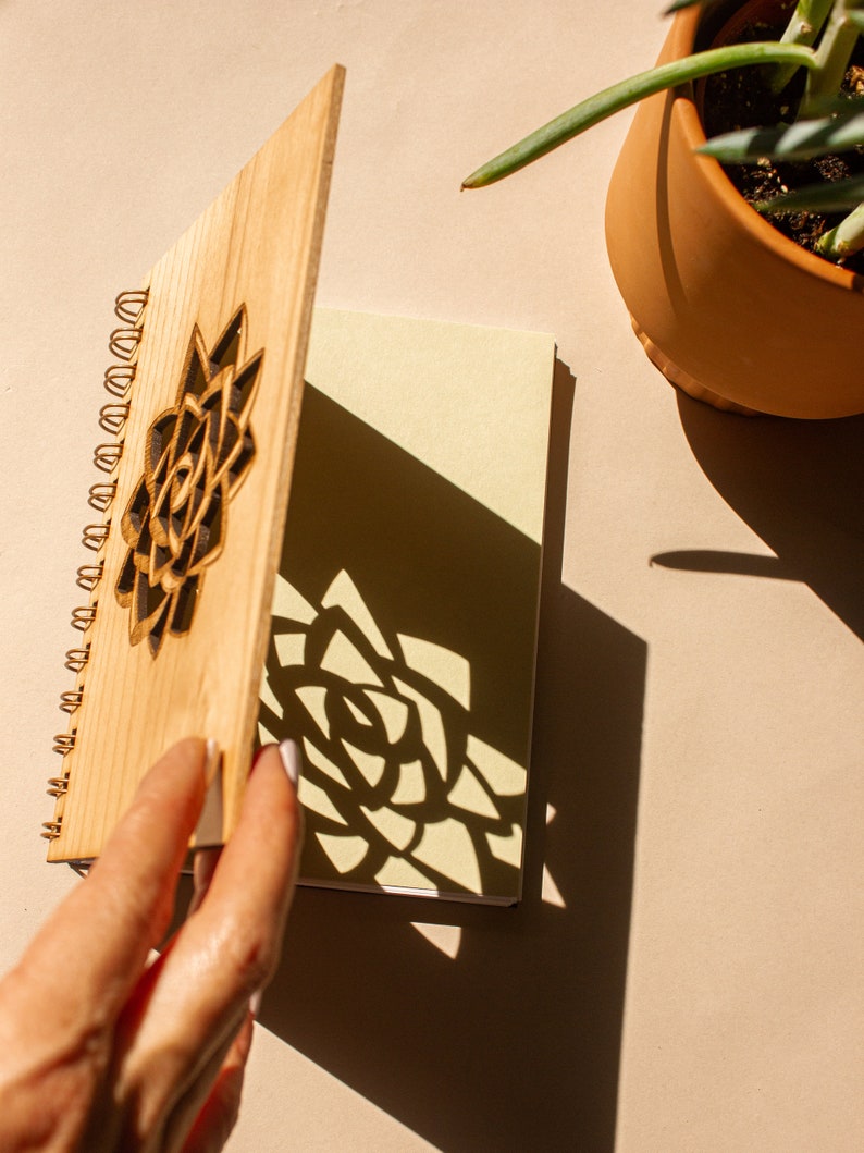 Puede incluir: Un cuaderno de madera con espiral y un dise&ntilde;o floral cortado con l&aacute;ser en la portada. El cuaderno est&aacute; abierto, revelando p&aacute;ginas en blanco. La sombra del dise&ntilde;o floral se proyecta sobre las p&aacute;ginas. Una planta en maceta est&aacute; en el fondo.
