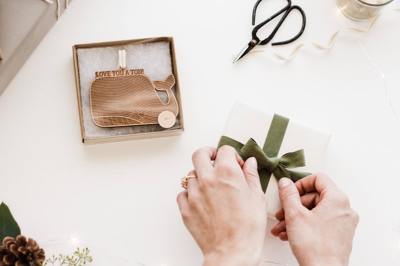 May include: A white gift box with a green ribbon tied in a bow. A wooden whale ornament with the text "Love you a ton!" is in the background.