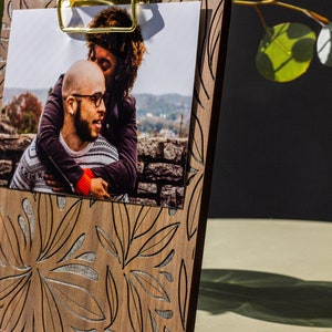 May include: A wooden photo frame with a gold clip and a floral design. The frame is holding a photo of a couple.
