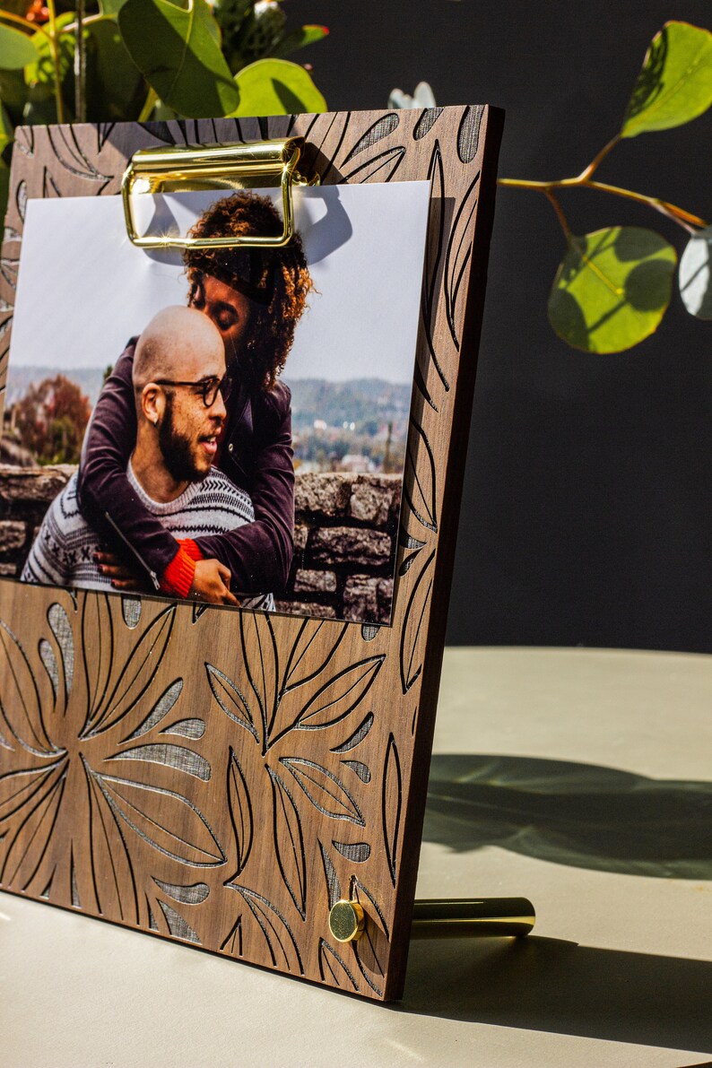 May include: A wooden photo frame with a gold clip and a floral design. The frame is holding a photo of a couple.