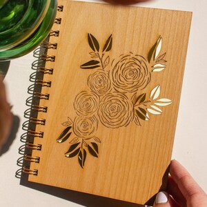 Puede incluir: Un cuaderno de espiral de madera con un dise&ntilde;o floral dorado. El dise&ntilde;o presenta un ramo de rosas y hojas.
