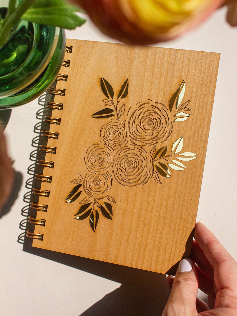 Puede incluir: Un cuaderno de espiral de madera con un dise&ntilde;o floral dorado. El dise&ntilde;o presenta un ramo de rosas y hojas.