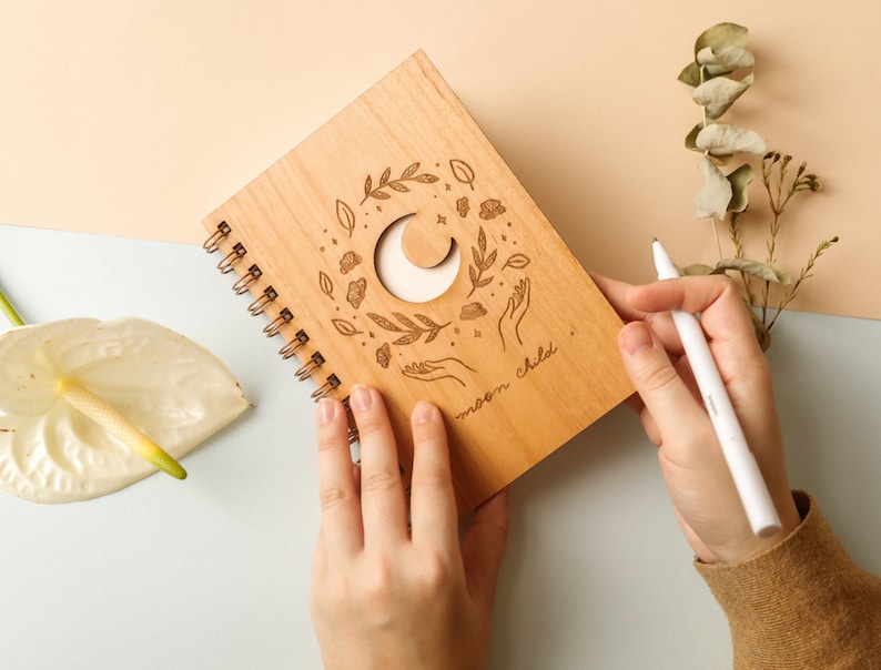 May include: A wooden journal with a spiral binding and a crescent moon cut-out design. The cover features an engraved design of leaves, stars, and hands reaching up to the moon. The text "moon child" is engraved on the cover.