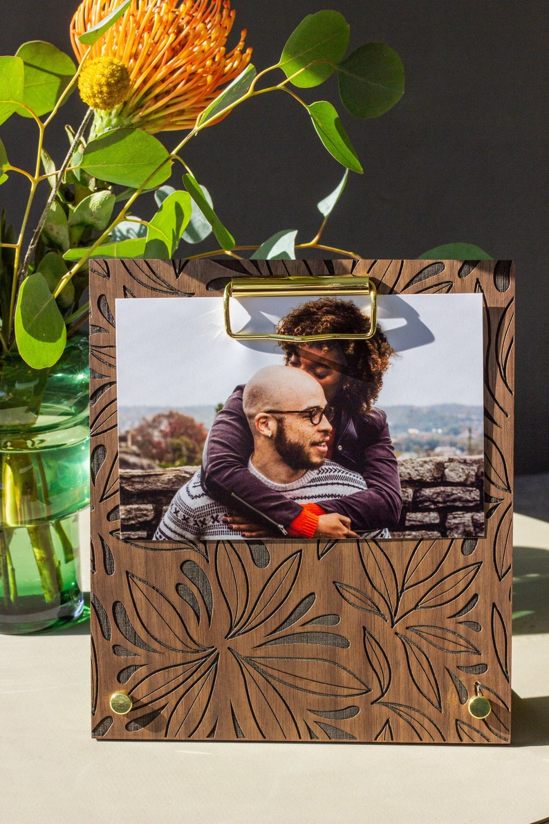 May include: A wooden photo frame with a floral design and a gold metal clip. The frame holds a photo of a couple embracing.