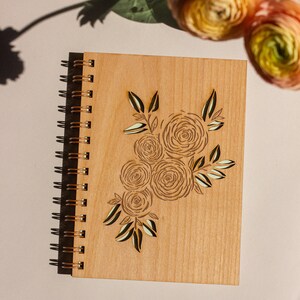 Puede incluir: Un diario de madera con encuadernaci&oacute;n en espiral y un dise&ntilde;o de ramo de flores cortado con l&aacute;ser. El dise&ntilde;o presenta varias rosas estilizadas y hojas de color marr&oacute;n claro.
