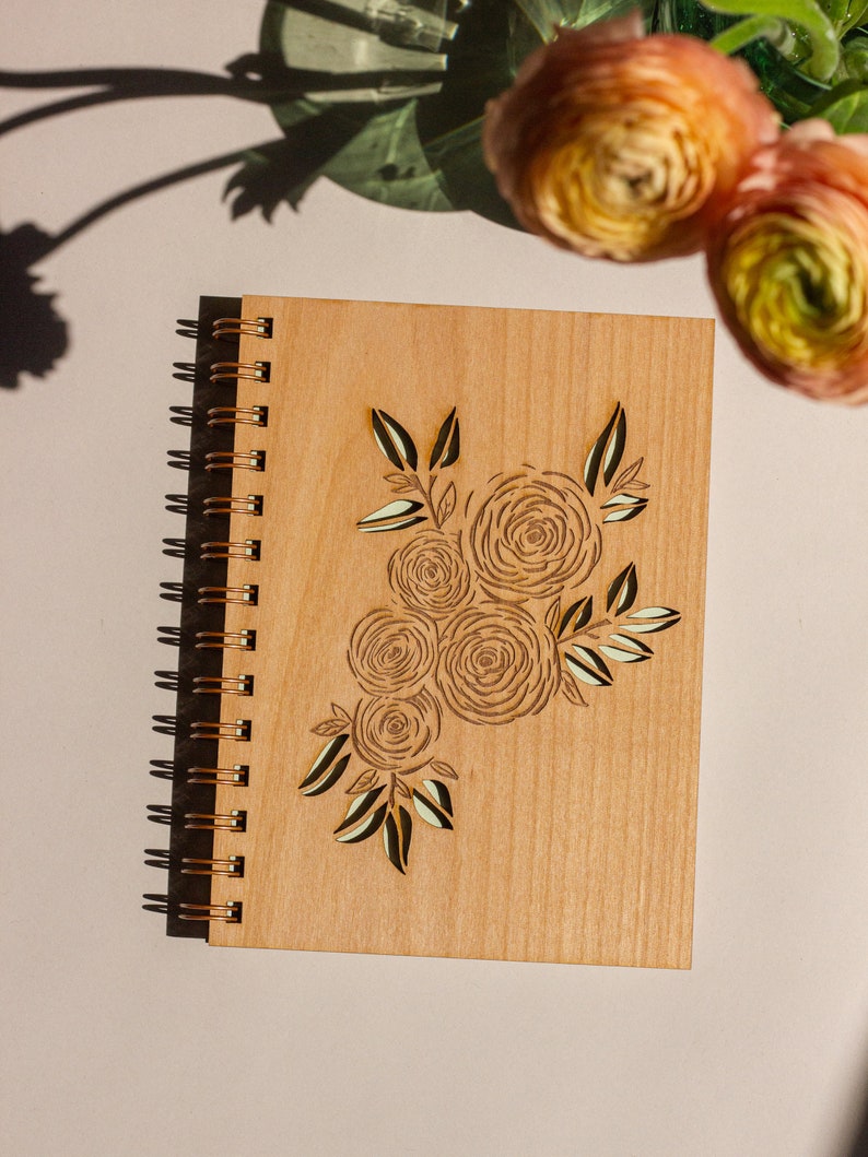 Puede incluir: Un diario de madera con encuadernaci&oacute;n en espiral y un dise&ntilde;o de ramo de flores cortado con l&aacute;ser. El dise&ntilde;o presenta varias rosas estilizadas y hojas de color marr&oacute;n claro.