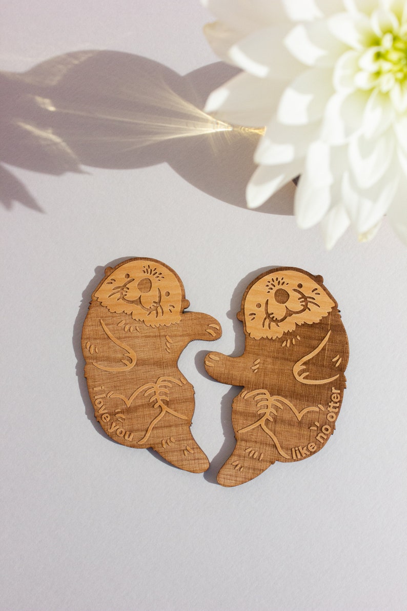 May include: Two wooden sea otter figurines holding each other. The otter on the left says "I love you" and the otter on the right says "Like no otter".