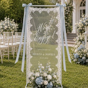 Puede incluir: Un marco rectangular de metal blanco que sostiene un panel de tela transparente con borde de encaje blanco. El panel presenta el texto "We're Engage!", "LET'S CELEBRATE!", "HANZO & SOPHIA" y "30.09.2026". Cintas de satén azul claro están atadas.
