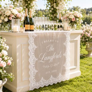 May include: A white lace table runner draped over a cream-colored table, featuring the printed text "CHEERS TO The Langfords EST. 2026" with a heart and leaf design. The runner has intricate scalloped lace edges and a sheer, semi-transparent center.