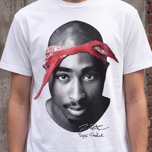 May include: A white short-sleeved t-shirt featuring a large black and white portrait of Tupac Shakur wearing a red bandana. Below the portrait is a black signature and the printed text Tupac Shakur. The design is centered on the chest area.
