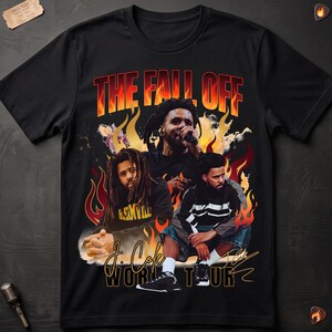 Könnte beinhalten: Ein schwarzes Kurzarm-T-Shirt mit einem Grafikdruck von J. Cole sowie dem Text THE FALL OFF und J. COLE WORLD TOUR. Das Design zeigt Flammen, drei Bilder des Künstlers und eine Unterschrift. Das T-Shirt liegt auf einer dunklen Oberfläche.