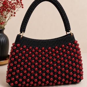 May include: A black crocheted handbag featuring a structured top handle and a body densely embellished with glossy, deep red spherical beads. The bag is attached to the handle with gold-toned metal rings, creating a distinct contrast between the dark fabric and the vibrant red accents.