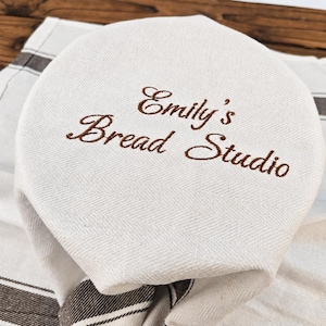 May include: A round, off-white fabric bread cover featuring a subtle herringbone weave pattern. The text "Emily's Bread Studio" is embroidered in brown thread across the centre. The cover is draped over a matching off-white cloth with brown stripes.