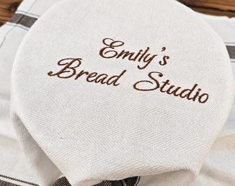 May include: A round, off-white fabric bread cover featuring a subtle herringbone weave pattern. The text "Emily's Bread Studio" is embroidered in brown thread across the centre. The cover is draped over a matching off-white cloth with brown stripes.
