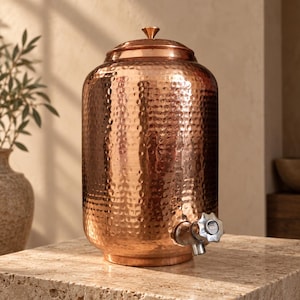 May include: A cylindrical copper water dispenser featuring a hammered texture across its surface. The vessel includes a matching domed lid with a small central knob and a silver-toned metal spigot attached to the lower front section for dispensing.