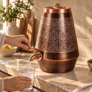 Puede incluir: Un dispensador de bebidas de cobre con cuerpo cilíndrico cónico que presenta un intrincado patrón de damasco oscuro grabado en la mitad superior, una tapa a juego con un pequeño pomo y un grifo metálico que vierte agua en un vaso.