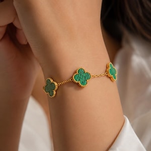 Könnte beinhalten: Ein goldenes Kettenarmband mit drei kleeblattförmigen Anhängern mit grünen Malachit-Steineinlagen. Jedes Kleeblatt ist mit einem perlenartigen Goldrand versehen, und die Anhänger sind durch eine feine Goldgliederkette miteinander verbunden.