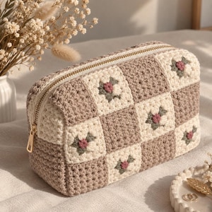May include: A rectangular crochet pouch featuring a checkered pattern of solid beige squares and cream squares with small embroidered pink flowers and green leaves. The pouch has a gold-toned metal zipper closure along the top edge.