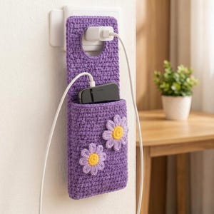 Könnte beinhalten: Ein violetter Handyhalter aus Häkelarbeit, der an einer weißen Steckdose hängt. Der Halter ist mit zwei Gänseblümchen-Applikationen mit hellvioletten Blütenblättern und gelben Mitten verziert. Ein schwarzes Smartphone steckt in der Tasche.