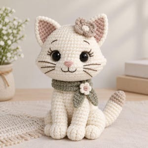 Puede incluir: Un juguete de peluche de gato tejido a ganchillo en color crema con grandes ojos negros, nariz rosa y bigotes bordados. El gato lleva una bufanda tejida de color verde salvia adornada con una pequeña flor blanca y una flor a juego en la oreja.