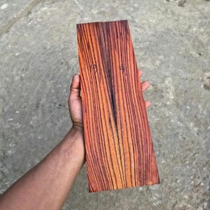 Può includere: Un pezzo di legno rettangolare con una venatura distinta caratterizzata da strisce verticali alternate in tonalità marrone rossastro intenso e arancione chiaro. La superficie presenta un segno scritto a mano con i numeri 19 e 19.