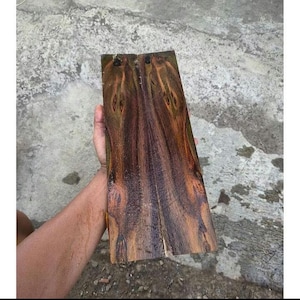Puede incluir: Una pieza rectangular de madera de color marrón oscuro con un patrón de veta vertical prominente que presenta vetas arremolinadas de color ámbar y negro. La superficie muestra un acabado natural pulido con nudos distintos y variaciones de color orgánicas.