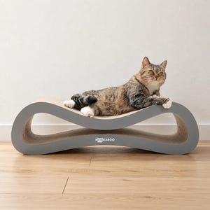 Puede incluir: Un rascador para gatos gris y curvado con una superficie de cartón texturizado y el texto blanco NOOKAROO impreso en el frente. Un gato atigrado marrón y negro con patas blancas está tumbado sobre el rascador, que tiene forma de infinito.