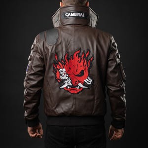 May include: A dark brown leather jacket featuring a large red and white graphic of a stylized demon mask on the back. The high collar displays the word SAMURAI in white text. The jacket includes black ribbed cuffs and hem, with circular accents on the sleeves.