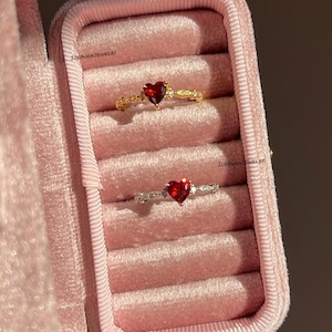 May include: Two rings with heart-shaped red stones are displayed in a pink velvet jewellery box. The top ring is gold-toned with a textured band, while the bottom ring is silver-toned with a smooth band. Both rings feature small clear accent stones.