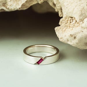 May include: A silver band ring featuring a single, rectangular, baguette-cut ruby gemstone set diagonally into the metal. The ring has a smooth, polished finish and a clean, modern design with a notched detail where the stone is embedded.