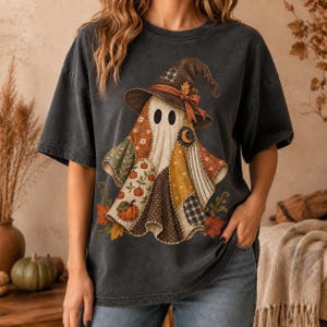May include: A charcoal gray short-sleeve t-shirt featuring a graphic of a ghost wearing a witch hat. The ghost is composed of various patchwork patterns including pumpkins, stars, and plaid, accented with autumn leaves and a crescent moon.