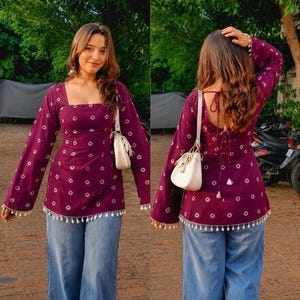 Könnte beinhalten: Eine dunkelviolette Tunika mit langen Ärmeln, weißem geometrischem Muster, quadratischem Ausschnitt und weißer Quastenborte am Saum. Das Kleidungsstück wird mit einer weiten blauen Jeans und einer kleinen weißen Umhängetasche mit Kordelzug kombiniert.