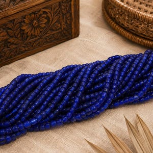 May include: A long, continuous strand of small, round, deep cobalt blue beads. The beads are uniform in size and shape, featuring a smooth, polished finish that reflects light. The strand is arranged in a dense, multi-layered coil across the frame.