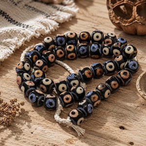 May include: A strand of black glass beads featuring circular eye-like patterns in shades of blue, tan, and white. The beads are strung on a thick, off-white braided cord with frayed ends, arranged in a loose coil on a textured wooden surface.