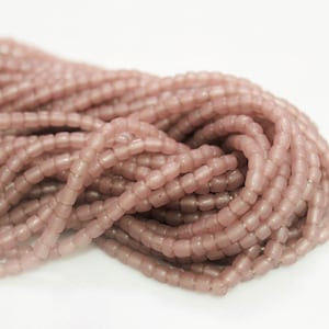 Puede incluir: Una hebra de cuentas de vidrio de color rosa polvoriento. Las cuentas son pequeñas y tienen una forma ligeramente redondeada y rectangular.